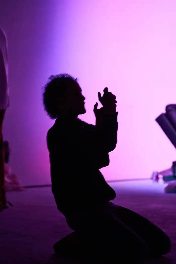 A participant kneeling with hands raised, silhouetted against projected violet light