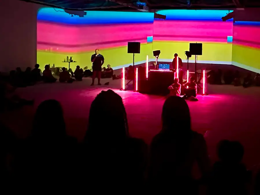 A facilitator addressing the room during the opening ceremony, the audience seated around a projected wall of colour