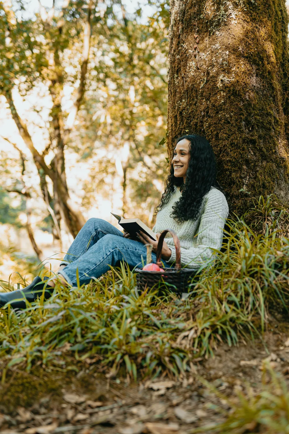 A woman reading against a mossy forest tree, a foraging basket beside her in the grass
